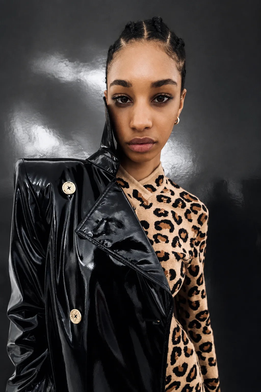 A woman wearing a glossy black vinyl trench coat over a leopard-print top against a dark metallic wall