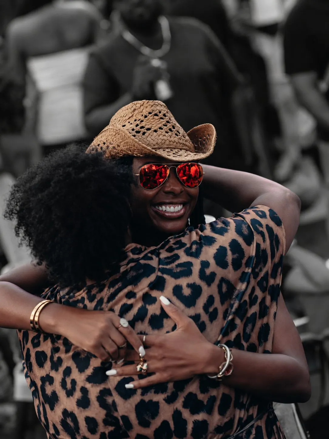 Two friends embracing at a festival, leopard print and red sunglasses in colour against a grey crowd