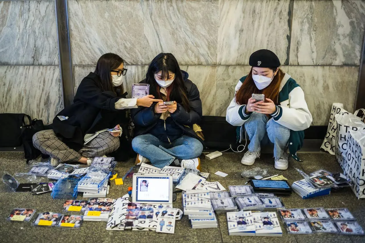 Three masked fans sit beside a stone wall with albums, photocards and open binders arranged in front of them