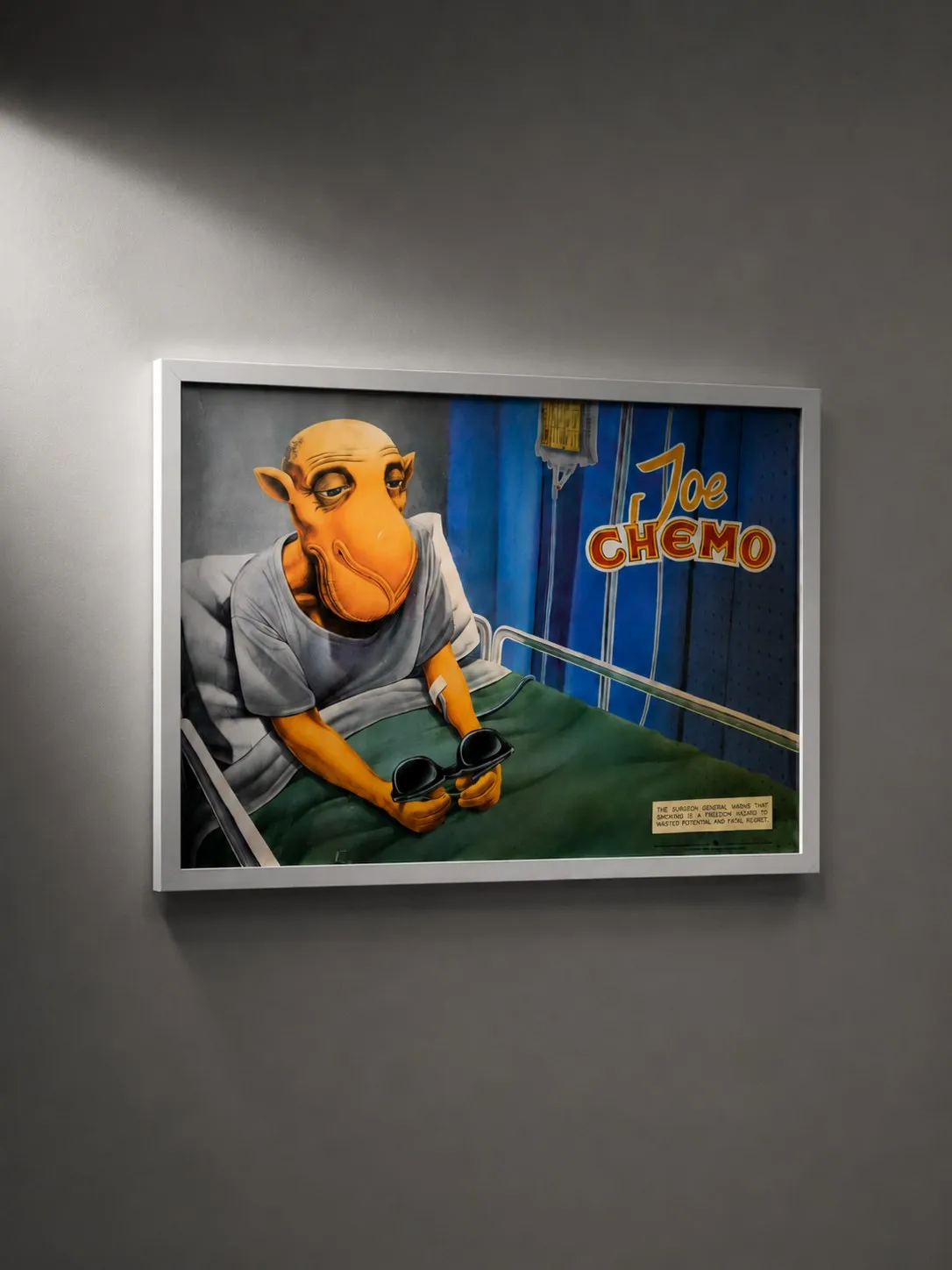 A framed Joe Chemo poster on a gallery wall, the orange camel lying in a hospital bed against a monochrome room