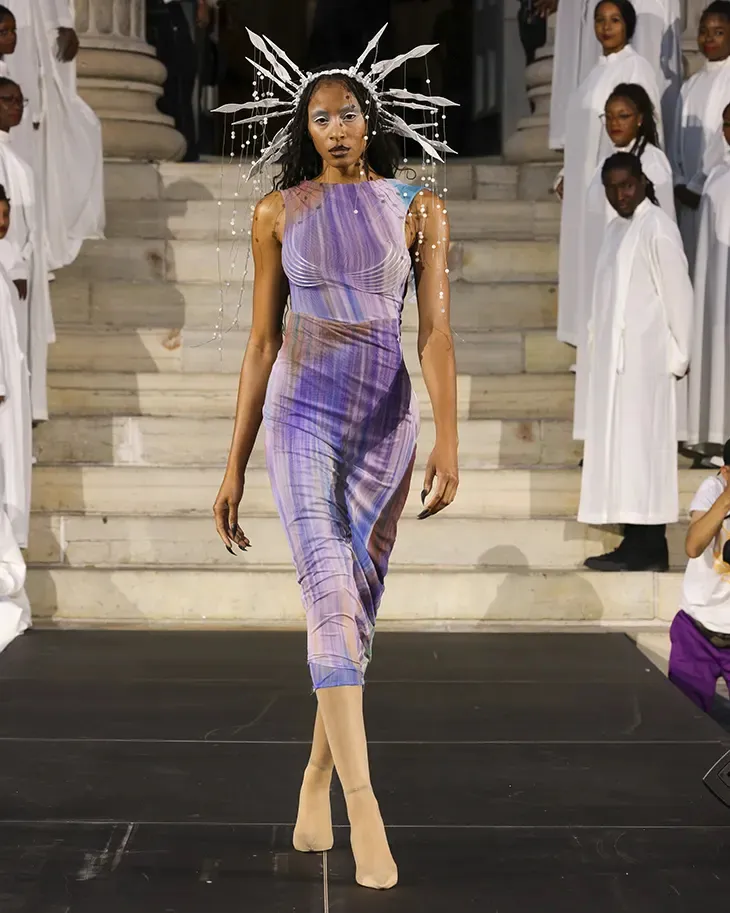 A model wearing a fitted purple dress and a radiating silver headpiece walks the runway on the steps of Brooklyn Borough Hall
